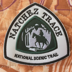 Może przedstawiać: Naszywka Natchez Trace National Scenic Trail. Naszywka jest trójkątna z białym obramowaniem i zawiera tekst "Natchez Trace" i "National Scenic Trail". Środek przedstawia jeźdźca na koniu w leśnej scenerii.