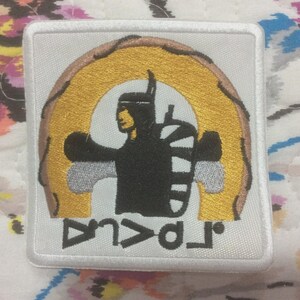 Könnte beinhalten: Quadratischer weißer Aufnäher mit einem gestickten Design. Das Design zeigt eine schwarze Silhouette einer Person mit ausgestreckten Armen, vor einem goldenen und braunen Bogen. Das Wort "AVALA" ist unten in Weiß gestickt.