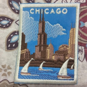 Può includere: Toppa ricamata raffigurante lo skyline di Chicago con barche a vela. La toppa ha uno sfondo blu con la scritta "CHICAGO" in alto. Gli edifici sono marroni e oro, e le barche a vela hanno vele bianche.