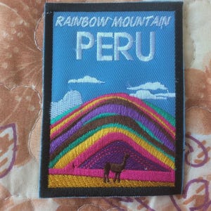 Może przedstawiać: Haftowana naszywka przedstawiająca Tęczową Górę w Peru. Naszywka ma niebieskie tło z białymi chmurami i napisem "RAINBOW MOUNTAIN PERU". Na dole znajduje się sylwetka lamy.