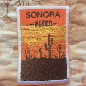 Puede incluir: Parche bordado con un paisaje desértico con cactus bajo un cielo al atardecer. El parche tiene el texto "SONORA - MEXICO" en letras marrones sobre un fondo naranja, enmarcado por un borde blanco.
