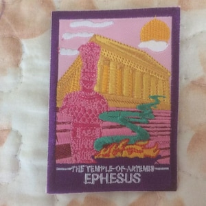 Può includere: Toppa ricamata raffigurante un tempio rosa con colonne gialle, un fiume verde e un sole giallo. Il testo "THE TEMPLE OF ARTEMIS EPHESUS" è in basso, incorniciato in viola.
