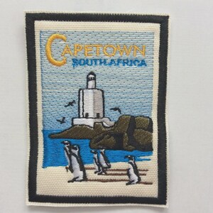 May include: Embroidered patch featuring a lighthouse, penguins, and the text "CAPETOWN SOUTH AFRICA." The design includes a blue ocean, brown rocks, and a light blue sky. The patch has a black border and a cream-colored background.