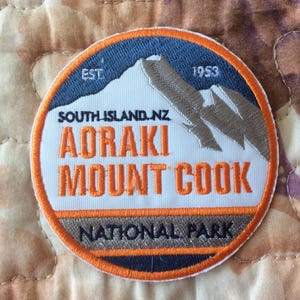 May include: Embroidered patch featuring the text "AORAKI MOUNT COOK NATIONAL PARK" with a mountain graphic. The patch is circular, with orange, blue, white, and gray colors. The text "SOUTH ISLAND.NZ" and the year "1953" are also included.