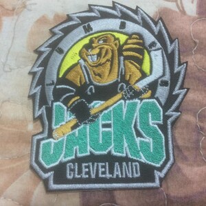 Könnte beinhalten: Gestickter Aufnäher mit einem Biber-Maskottchen, das einen Hockeyschläger hält, vor einem Sägeblatt-Design. Der Aufnäher enthält das Wort "Jacks" in Grün und die Stadt "Cleveland" in Weiß.