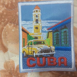May include: Embroidered patch featuring a yellow vintage car in front of a tall yellow building with a red-tiled roof. The word "CUBA" is stitched in red letters at the bottom, set against a blue background.
