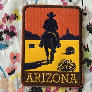 Puede incluir: Parche bordado con un vaquero a caballo silueteado contra un atardecer naranja. El parche tiene un borde marrón y la palabra "ARIZONA" en letras doradas. El fondo es amarillo y azul.