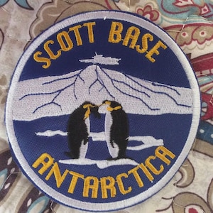 May include: Embroidered patch with a blue background featuring the text "SCOTT BASE ANTARCTICA" in yellow. The patch depicts a mountain range, a building, and two penguins. The patch has a white border.
