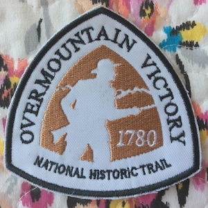 Może przedstawiać: Biało-brązowa naszywka z napisem "OVERMOUNTAIN VICTORY" i "NATIONAL HISTORIC TRAIL". Naszywka przedstawia sylwetkę osoby i rok 1780.