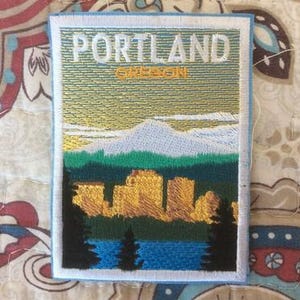 May include: Embroidered patch featuring a landscape scene with the text "PORTLAND OREGON". The design includes a mountain range, trees, and a body of water, with a blue border.