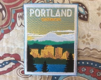 May include: Embroidered patch featuring a landscape scene with the text "PORTLAND OREGON". The design includes a mountain range, trees, and a body of water, with a blue border.