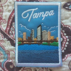 Könnte beinhalten: Gestickter Aufnäher mit dem Wort "Tampa" in weißer Schrift über einer Skyline. Der Aufnäher zeigt einen blauen Himmel mit Wolken, eine Uferpromenade und Gebäude. Der rechteckige Aufnäher hat einen weißen Rand.