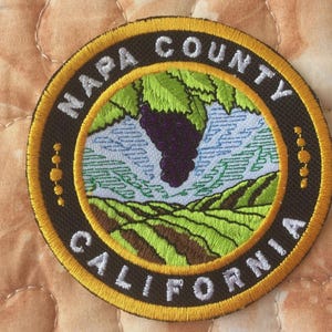 May include: Embroidered patch featuring the words "NAPA COUNTY CALIFORNIA" in white lettering around a circular design. The design includes a cluster of purple grapes, green leaves, and rolling green hills. The patch has a black background with a gold border.