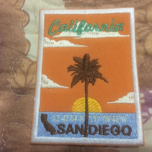 Peut inclure: Écusson brodé représentant un coucher de soleil californien avec un palmier, un soleil et des nuages. L'écusson comprend le texte "California" et "San Diego" avec des coordonnées. L'écusson a une bordure blanche.