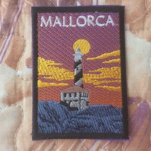 Può includere: Toppa ricamata raffigurante una scena di faro con la scritta "MALLORCA" in alto. Il disegno comprende un faro a strisce bianche e nere, un sole giallo e un cielo al tramonto con tonalità arancioni e viola. La toppa è rettangolare con un bordo nero.