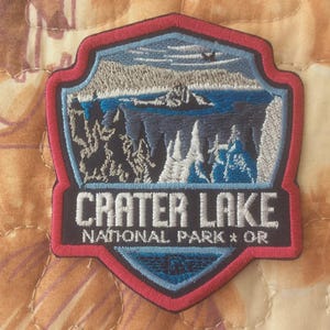 May include: Embroidered patch featuring a scenic view of Crater Lake National Park in Oregon. The patch has a shield shape with a red border, depicting a lake, mountains, and trees in shades of blue, gray, and white. The text "CRATER LAKE NATIONAL PARK * OR" is embroidered in white.