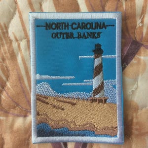 May include: Embroidered patch featuring a lighthouse with brown and white stripes, set against a blue sky and sandy beach. The text "NORTH CAROLINA OUTER BANKS" is embroidered at the top. The patch has a white border.