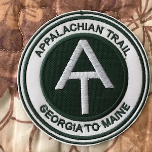 May include: Circular patch with the text "APPALACHIAN TRAIL GEORGIA TO MAINE" in white lettering. The center features a white "A" and "T" symbol on a dark green background, surrounded by a white border.