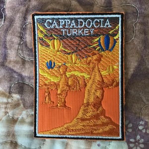 Op de afbeelding: Een rechthoekige geborduurde patch met de tekst CAPPADOCIA TURKEY bovenaan. Het ontwerp toont een landschap met oranje rotsformaties en verschillende kleurrijke heteluchtballonnen die in een gele en oranje lucht zweven, met een zwarte rand.