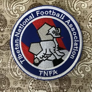 May include: A circular embroidered patch featuring a blue border with the white text Tibetan National Football Association and TNFA. The centre displays a stylised silver snow lion graphic over a red diagonal stripe on a blue background.