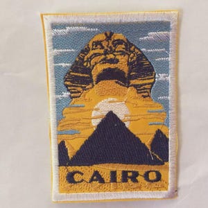 Może przedstawiać: Haftowana naszywka przedstawiająca Sfinksa, piramidy i wschodzące słońce na tle błękitnego nieba. Słowo "CAIRO" jest wyświetlane pogrubioną żółtą czcionką u dołu. Naszywka ma białą obwódkę.