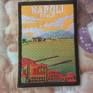 Könnte beinhalten: Gestickter Aufnäher mit einer Landschaftsszene und dem Text "NAPOLI ITALY". Der Aufnäher hat einen gelben Himmel, weiße Wolken, grüne Felder und rote Gebäude. Er ist mit einem schwarzen Rand eingefasst.