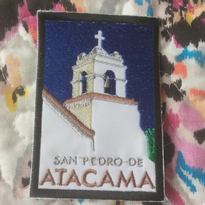 May include: Embroidered patch featuring a church tower with a cross on top, set against a blue sky. The patch reads "SAN PEDRO-DE-ATACAMA" in white lettering, framed by a black border. The patch is rectangular.