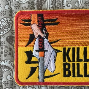 May include: A square embroidered patch featuring a yellow and orange gradient background with black Japanese kanji characters. The design depicts a hand holding a sword wrapped in a white cloth, with the black text KILL BILL in the bottom right.