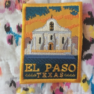 May include: Embroidered patch depicting a church in El Paso, Texas. The patch has a yellow border and includes the text "EL PASO TEXAS" in a dark blue banner. The background shows a sunset with orange and blue hues. The patch is rectangular.