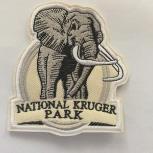 Könnte beinhalten: Gestickter Aufnäher mit einem grauen Elefanten mit Stoßzähnen auf cremefarbenem Hintergrund. Der Text "NATIONAL KRUGER PARK" ist in schwarzer Schrift mit weißer und grauer Umrandung dargestellt. Ein Souvenir-Patch.