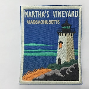 Puede incluir: Parche bordado con un faro en una costa rocosa, con el texto "MARTHA'S VINEYARD MASSACHUSETTS". El faro es blanco con la parte superior negra y una ventana naranja, sobre un cielo azul y mar.