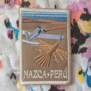 Può includere: Toppa ricamata raffigurante le Linee di Nazca in Perù. Il disegno include un uccello, un aereo e la scritta "NAZCA PERÚ" in bianco. La toppa ha uno sfondo beige con un bordo bianco.