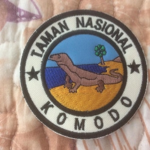 May include: Circular patch with the text "TAMAN NASIONAL KOMODO" in white on a black border. The patch features a Komodo dragon illustration on a beach with a tree and blue sky.