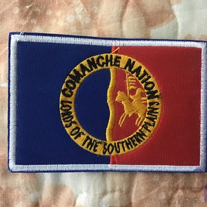 May include: A rectangular embroidered patch featuring a vertical split design with a dark blue left side and a red right side. A yellow circular border contains the black text COMANCHE NATION LORDS OF THE SOUTHERN PLAINS and a yellow horse icon.