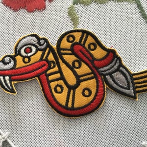 Puede incluir: Un parche bordado con un diseño de serpiente estilizada en hilos de color amarillo, rojo, negro y plateado. La serpiente tiene una boca roja prominente, colmillos blancos y una cabeza y cola plateadas, todo delineado con un borde negro grueso.