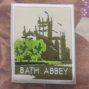 Op de afbeelding: Geborduurde patch met Bath Abbey, een historische kerk, met groene bomen en de tekst "BATH ABBEY". De patch heeft een witte rand en staat op een beige achtergrond.