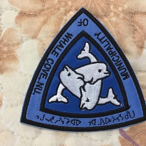 May include: A triangular embroidered patch with a blue background and a black border. The center features three white beluga whales arranged in a circular pattern. The text MUNICIPALITY OF WHALE COVE, NU is embroidered in black along the edges.