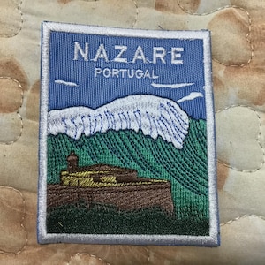 May include: Embroidered patch featuring the text "NAZARE PORTUGAL" in white on a blue background. The patch depicts a coastal scene with a mountain range, green hills, and a building. The patch has a white border.