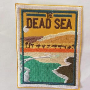 Puede incluir: Parche bordado con un paisaje del Mar Muerto. El diseño incluye el texto "THE DEAD SEA" en marrón sobre un fondo amarillo. El parche tiene forma rectangular con un borde blanco y un borde exterior amarillo.