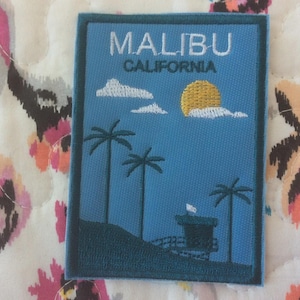 Może przedstawiać: Prostokątna naszywka z niebieskim tłem z białym napisem "MALIBU CALIFORNIA". Naszywka przedstawia palmy, żółte słońce, białe chmury i wieżę ratowniczą. Naszywka ma ciemnoniebieską obwódkę.