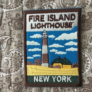 Puede incluir: Un parche bordado rectangular que muestra el faro de Fire Island en Nueva York. El diseño presenta un faro a rayas marrones y blancas, un edificio con techo verde y un campo amarillo bajo un cielo azul con nubes blancas y bordes marrones.