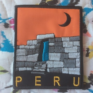 Op de afbeelding: Geborduurde patch met een Peruaans landschap met een stenen structuur, een blauwe deuropening en een halve maan tegen een oranje lucht. Het woord "PERU" is in het geel geborduurd aan de onderkant. De patch is rechthoekig.