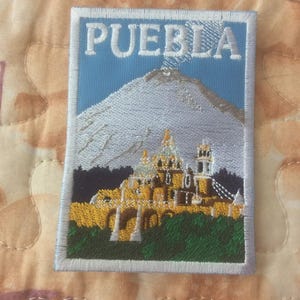 Op de afbeelding: Geborduurde patch met het woord "PUEBLA" boven een berg en een geel gebouw met torens. De patch heeft een blauwe hemelachtergrond en een witte rand. Het gebouw is geel en groen.