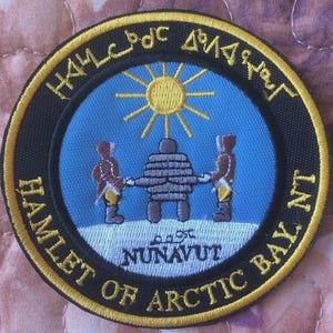 Könnte beinhalten: Runder, gestickter Aufnäher mit einem blauen Mittelteil, der einen Inukshuk, zwei Figuren und eine Sonne zeigt. Der Aufnäher hat Text in einem halbkreisförmigen Muster am Rand, mit einem schwarzen Rand und gelben Akzenten. Der Text lautet "HAMLET OF ARCTIC BAY, NU".