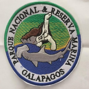 May include: Embroidered patch featuring a turtle and hammerhead shark, with text reading "PARQUE NACIONAL & RESERVA MARINA GALAPAGOS." The patch has a white border, green, blue, and brown colors.