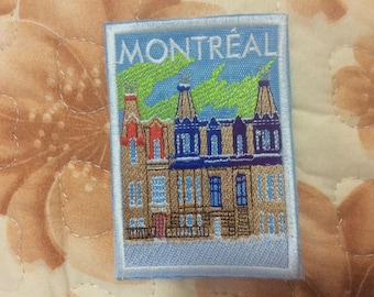 May include: Embroidered patch featuring a Montreal cityscape with the city name at the top. The patch depicts colorful buildings with green accents against a blue sky. The patch has a light blue border.