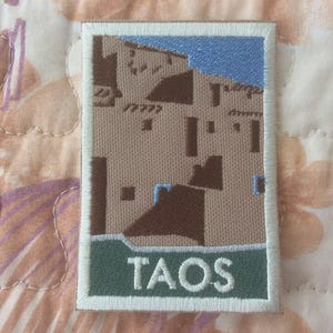 Puede incluir: Parche bordado con un diseño arquitectónico del suroeste con un fondo de cielo azul. El parche tiene un borde blanco y una pancarta verde en la parte inferior con la palabra "TAOS" en letras blancas.