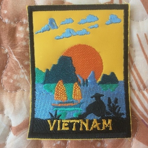 May include: Embroidered patch featuring a Vietnam landscape with a yellow sky, orange sun, and blue clouds. The design includes mountains, water, and a boat, with the word "VIETNAM" at the bottom.