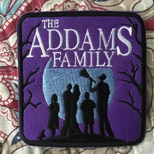 Puede incluir: Un parche bordado cuadrado con fondo morado y siluetas negras de una familia frente a una gran luna azul claro. El texto THE ADDAMS FAMILY está bordado en blanco en la parte superior, con ramas de árboles negras desnudas a los lados.