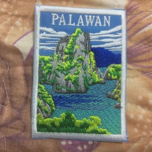 Pode incluir: Patch bordado com a palavra "PALAWAN" acima de uma paisagem detalhada de uma ilha tropical com vegetação verde exuberante, formações rochosas cinzentas e água azul. O patch tem uma borda branca.